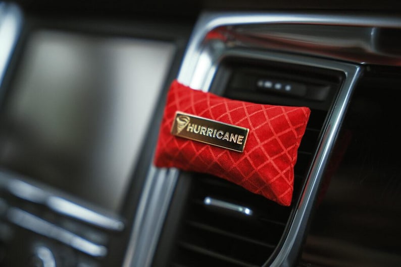 May include: A red, diamond-patterned air freshener with a gold-colored rectangular label that reads "HURRICANE". The air freshener is attached to a car's air vent, with a blurred background.