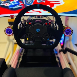 May include: A black racing simulator steering wheel with blue detailing and various buttons. The wheel is positioned in front of a racing seat, with a screen showing a race car in the background. The setup includes colourful lighting.