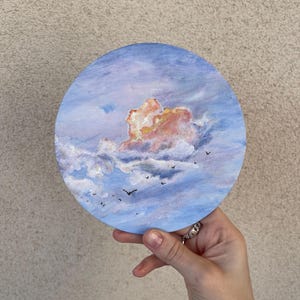 Könnte beinhalten: Rundes Gemälde eines bewölkten Himmels mit Vögeln. Das Kunstwerk zeigt eine große, flauschige Wolke in Rosa- und Orangetönen vor einem blau-weißen Himmel. Kleine schwarze Vögel sind über die Leinwand gemalt. Das Gemälde wird von einer Hand gehalten.