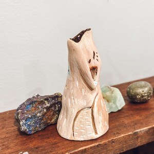 May include: A handmade, beige ceramic sculpture with a textured surface and carved details. The sculpture has a unique, abstract shape with a dark brown interior. It is displayed on a wooden shelf with colorful rocks.