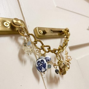 May include: A gold-coloured chain door lock with decorative charms. Charms include a horse, blue and white floral ceramic pendant, small bells, and beads. The lock is attached to a gold-coloured door handle on a white door.