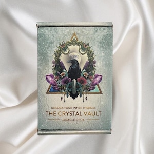 Könnte beinhalten: Rechteckige Schachtel für ein Orakeldeck. Die Schachtel zeigt ein Design mit einer schwarzen Krähe, lila Kristallen und floralen Elementen. Der Text auf der Schachtel lautet "UNLOCK YOUR INNER WISDOM: THE CRYSTAL VAULT ORACLE DECK."