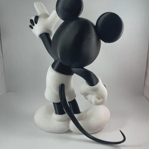 May include: A black, white, and gray figurine of Mickey Mouse, a classic cartoon character. The figure is posed with one arm raised, wearing white gloves, shorts, and shoes. The figure has large black ears and a long, curved tail.