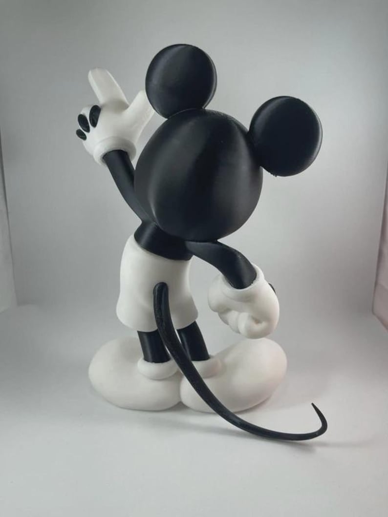 May include: A black, white, and gray figurine of Mickey Mouse, a classic cartoon character. The figure is posed with one arm raised, wearing white gloves, shorts, and shoes. The figure has large black ears and a long, curved tail.
