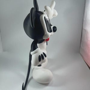 May include: A black and white cartoon character figurine with a red bow tie. The figure has large white gloves and shoes, with a black tail and ears. The character is posed with one hand raised.