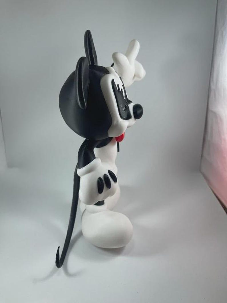 May include: A black and white cartoon character figurine with a red bow tie. The figure has large white gloves and shoes, with a black tail and ears. The character is posed with one hand raised.