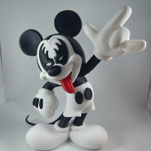 May include: A black, white, and red figurine of Mickey Mouse in a rock and roll pose. The figurine has a black and white face design, a red tongue, and white gloves. The figure is wearing white shorts and large white shoes.