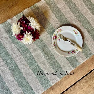 May include: A table runner with vertical sage green and beige stripes, featuring a bouquet of white and deep red chrysanthemums in a bowl, and a white ceramic plate with a floral rim pattern and a silver fork. Text reads: Handmade in Kent.