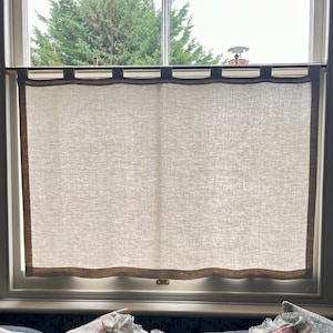 May include: A rectangular cafe curtain made of light beige, semi-sheer fabric with a visible woven texture. The curtain features a darker brown border along the bottom and sides, and is suspended from a thin metal rod by several fabric loops.