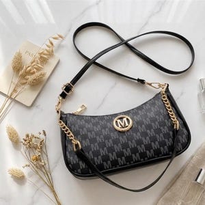 Könnte beinhalten: Schwarze und graue Umhängetasche mit sich wiederholendem "M"-Muster. Die Tasche hat einen goldenen Kettenriemen, ein goldenes "M"-Emblem und einen schwarzen, verstellbaren Schulterriemen. Ein Reisepass und Trockenblumen sind im Hintergrund.