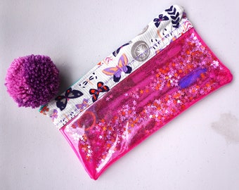 May include: A pink clear pencil case with a white fabric flap featuring a butterfly and floral design. The case is filled with pink glitter and stars.