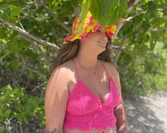 May include: A woman wearing a pink crochet top with a yellow and pink crocheted hat and blue denim shorts.