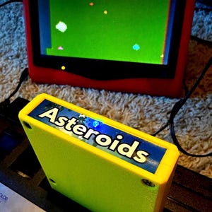 Peut inclure: Une cartouche de jeu vid&eacute;o jaune vif &eacute;tiquet&eacute;e Asteroids ins&eacute;r&eacute;e dans une console noire, plac&eacute;e devant un &eacute;cran rouge affichant un &eacute;cran vert avec le jeu Asteroids montrant un score de 140 et un nombre de vies de 1.