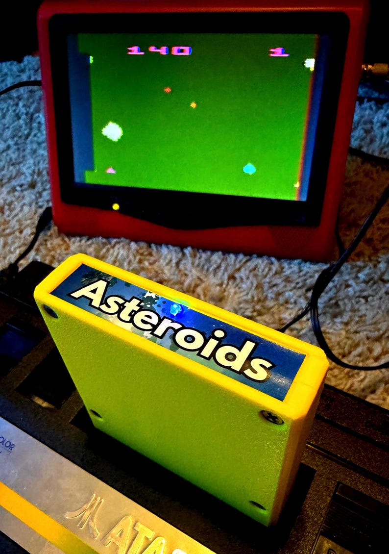 Peut inclure: Une cartouche de jeu vid&eacute;o jaune vif &eacute;tiquet&eacute;e Asteroids ins&eacute;r&eacute;e dans une console noire, plac&eacute;e devant un &eacute;cran rouge affichant un &eacute;cran vert avec le jeu Asteroids montrant un score de 140 et un nombre de vies de 1.