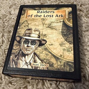 May include: A black Atari 2600 video game cartridge for Raiders of the Lost Ark. The label features an illustration of a person wearing a fedora and sunglasses over a vintage map background, with the game title and console name printed.