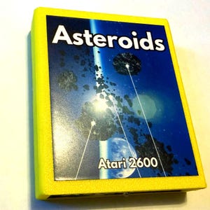 Peut inclure: Un bo&icirc;tier de cartouche de jeu vid&eacute;o Atari 2600 rectangulaire avec un cadre en plastique jaune vif. L'&eacute;tiquette centrale pr&eacute;sente une sc&egrave;ne spatiale bleu fonc&eacute; avec des ast&eacute;ro&iuml;des, une plan&egrave;te et le texte blanc Asteroids et Atari 2600.
