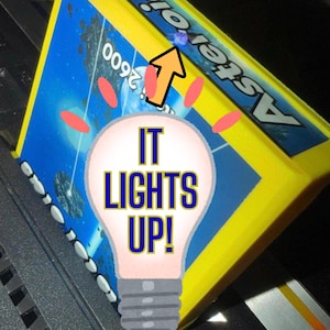May include: A yellow Atari 2600 Asteroids video game cartridge featuring a blue space-themed label with the text Asteroids and 2600. A lightbulb graphic with the text IT LIGHTS UP is overlaid on the center with an orange arrow pointing up.