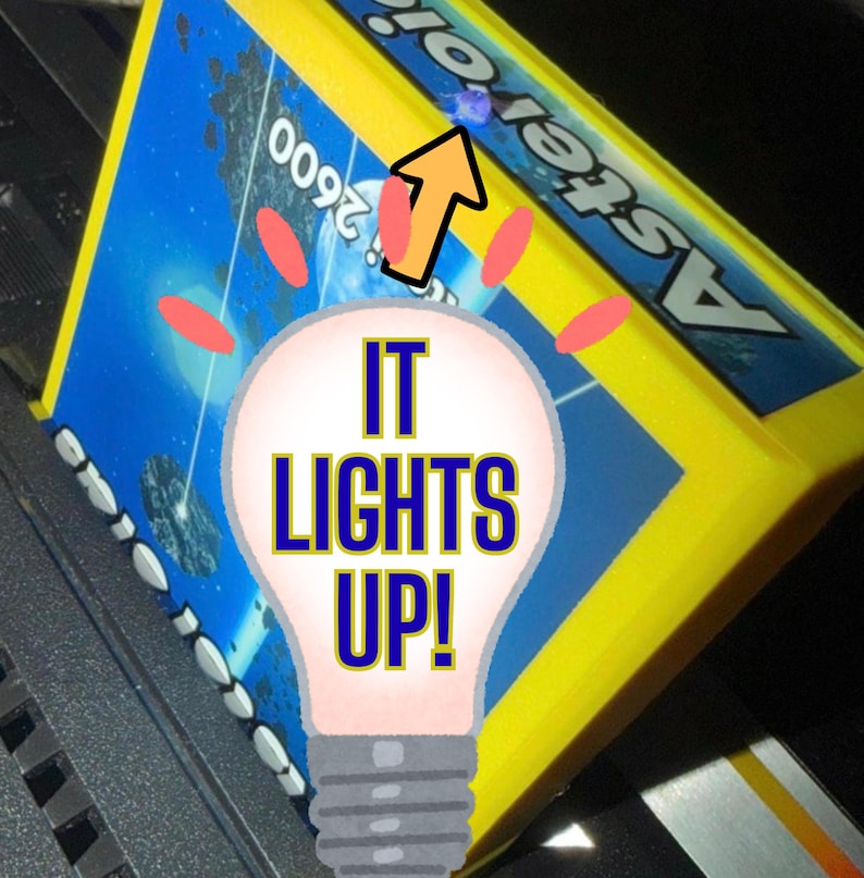 Peut inclure: Une cartouche de jeu vid&eacute;o Atari 2600 Asteroids jaune avec une &eacute;tiquette bleue sur le th&egrave;me de l'espace portant le texte Asteroids et 2600. Un graphique d'ampoule avec le texte IT LIGHTS UP est superpos&eacute; au centre avec une fl&egrave;che orange.