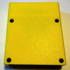 Peut inclure: Un bo&icirc;tier rectangulaire en plastique jaune avec une surface textur&eacute;e. L'objet pr&eacute;sente quatre trous circulaires situ&eacute;s pr&egrave;s de chaque coin, avec une seule vis noire visible dans le trou en bas &agrave; droite, tandis que les trois autres trous semblent vides.