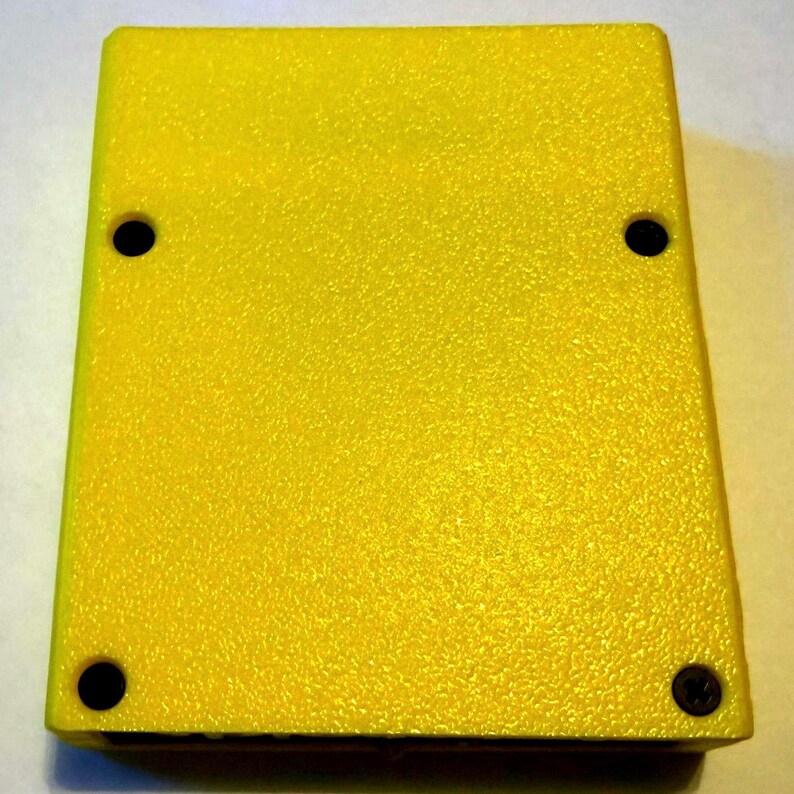 Peut inclure: Un bo&icirc;tier rectangulaire en plastique jaune avec une surface textur&eacute;e. L'objet pr&eacute;sente quatre trous circulaires situ&eacute;s pr&egrave;s de chaque coin, avec une seule vis noire visible dans le trou en bas &agrave; droite, tandis que les trois autres trous semblent vides.