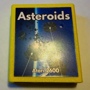 Peut inclure: Une cartouche de jeu vid&eacute;o Atari 2600 rectangulaire et jaune pour le jeu Asteroids. L'&eacute;tiquette centrale pr&eacute;sente un fond spatial bleu avec des ast&eacute;ro&iuml;des sombres, des lignes laser blanches et le texte Asteroids et Atari 2600 en blanc.