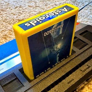 Peut inclure: Une cartouche de jeu vid&eacute;o Atari 2600 jaune pour le jeu Asteroids ins&eacute;r&eacute;e dans une console noire. L'&eacute;tiquette de la cartouche pr&eacute;sente une sc&egrave;ne spatiale bleu fonc&eacute; avec le texte blanc Asteroids et Atari 2600. Une petite lumi&egrave;re bleue est visible.