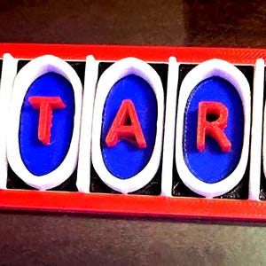 May include: A rectangular red base supports five white oval frames, each containing a blue oval insert. Centered on each blue insert is a red letter, collectively spelling the word ATARI. The structure is composed of distinct, layered components.