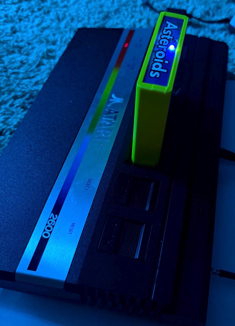 Peut inclure: Une console de jeux vid&eacute;o Atari 2600 noire avec un panneau de commande argent&eacute; portant le logo Atari, les mentions Select et Reset. Une cartouche de jeu Asteroids vert citron vif est ins&eacute;r&eacute;e dans la fente sup&eacute;rieure.