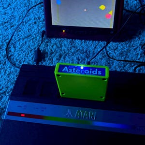 Peut inclure: Une console de jeux vid&eacute;o Atari 2600 noire avec une cartouche de jeu Asteroids vert vif ins&eacute;r&eacute;e. Un petit &eacute;cran au-dessus affiche le jeu en cours avec des formes g&eacute;om&eacute;triques color&eacute;es sur un fond sombre sous un &eacute;clairage bleu.