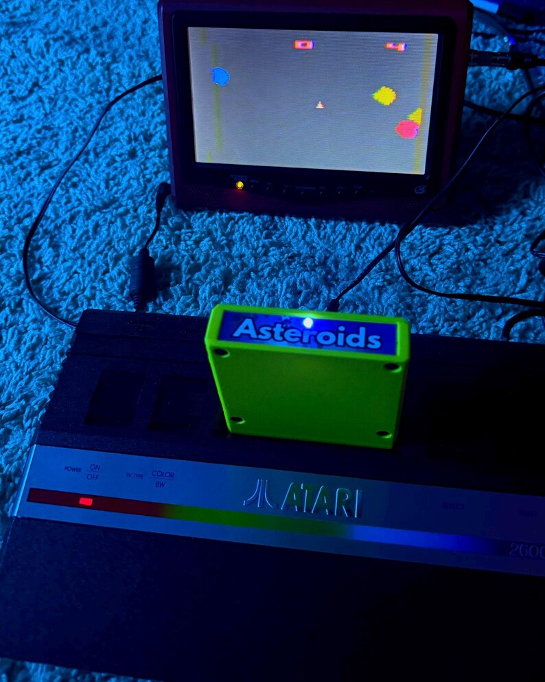 Peut inclure: Une console de jeux vid&eacute;o Atari 2600 noire avec une cartouche de jeu Asteroids vert vif ins&eacute;r&eacute;e. Un petit &eacute;cran au-dessus affiche le jeu en cours avec des formes g&eacute;om&eacute;triques color&eacute;es sur un fond sombre sous un &eacute;clairage bleu.