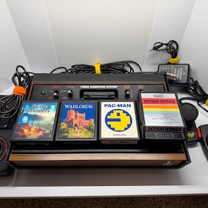 May include: An Atari Video Computer System console with wood-grain trim, two black joystick controllers, and two paddle controllers. Four game cartridges are included: Space Invaders, Warlords, Pac-Man, and Demon Attack, along with power cables.