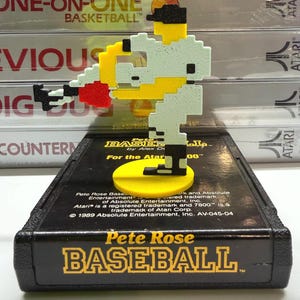 May include: A 3D-printed pixelated baseball player figure in white, yellow, black, and red, standing on a yellow circular base atop a black Pete Rose Baseball Atari 7800 game cartridge. The cartridge features yellow text reading Pete Rose BASEBALL.