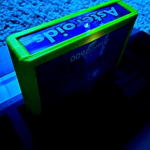 Peut inclure: Une cartouche de jeu vid&eacute;o rectangulaire Atari 2600 pour le jeu Asteroids. La cartouche pr&eacute;sente un bo&icirc;tier en plastique vert citron vif entourant une &eacute;tiquette bleue avec le texte blanc Asteroids et Atari 2600 imprim&eacute; dessus.