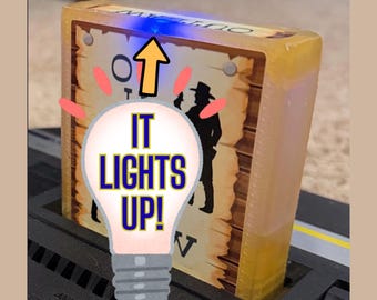 Peut inclure: Une cartouche de jeu vidéo rectangulaire avec un boîtier jaune translucide insérée dans une console noire. L'étiquette présente la silhouette d'un cow-boy et le texte "OUTLAW". Un graphique d'ampoule avec le texte "IT LIGHTS UP!" est superposé.