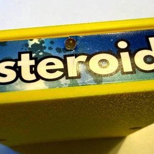 Peut inclure: Une cartouche de jeu vid&eacute;o rectangulaire jaune vif avec une &eacute;tiquette bleue portant le mot Asteroids imprim&eacute; en grandes lettres blanches. L'&eacute;tiquette comprend une petite lumi&egrave;re circulaire int&eacute;gr&eacute;e au centre au-dessus du texte.