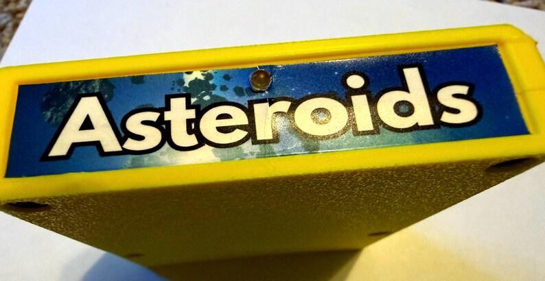 Peut inclure: Une cartouche de jeu vid&eacute;o rectangulaire jaune vif avec une &eacute;tiquette bleue portant le mot Asteroids imprim&eacute; en grandes lettres blanches. L'&eacute;tiquette comprend une petite lumi&egrave;re circulaire int&eacute;gr&eacute;e au centre au-dessus du texte.