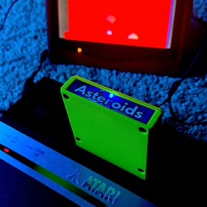 Peut inclure: Une cartouche de jeu Atari vert n&eacute;on vif &eacute;tiquet&eacute;e Asteroids, plac&eacute;e devant une console Atari noire et un &eacute;cran de t&eacute;l&eacute;vision affichant une interface de jeu rouge. La cartouche pr&eacute;sente une &eacute;tiquette bleue avec du texte blanc.