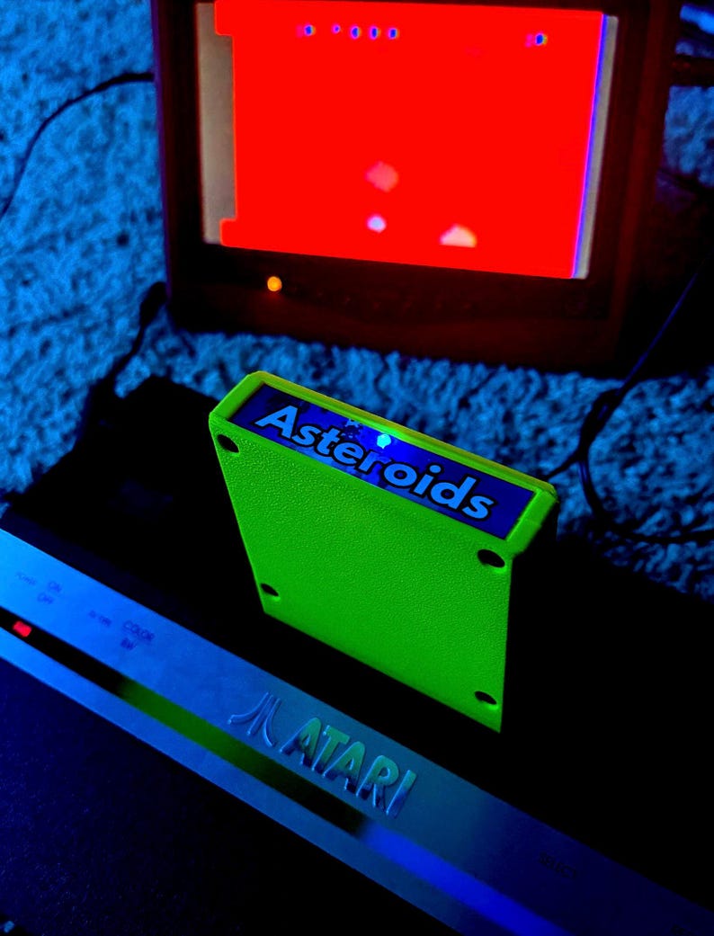 Peut inclure: Une cartouche de jeu Atari vert n&eacute;on vif &eacute;tiquet&eacute;e Asteroids, plac&eacute;e devant une console Atari noire et un &eacute;cran de t&eacute;l&eacute;vision affichant une interface de jeu rouge. La cartouche pr&eacute;sente une &eacute;tiquette bleue avec du texte blanc.