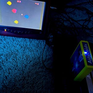 Peut inclure: Un petit &eacute;cran de t&eacute;l&eacute;vision affiche un jeu vid&eacute;o r&eacute;tro avec des formes pixelis&eacute;es color&eacute;es et un vaisseau triangulaire central. &Agrave; c&ocirc;t&eacute;, une cartouche vert vif &eacute;tiquet&eacute;e Asteroids pr&eacute;sente une lumi&egrave;re bleue brillante sur le dessus.