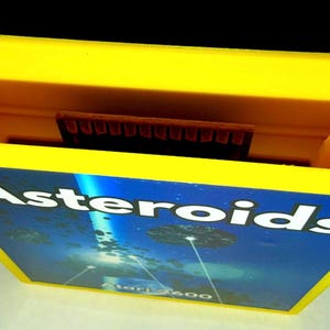 Peut inclure: Une cartouche de jeu vid&eacute;o Atari 2600 jaune vif pour le jeu Asteroids. L'&eacute;tiquette pr&eacute;sente le texte blanc Asteroids sur un fond spatial bleu fonc&eacute; avec des rochers flottants et un faisceau lumineux, avec Atari 2600 imprim&eacute; en dessous.