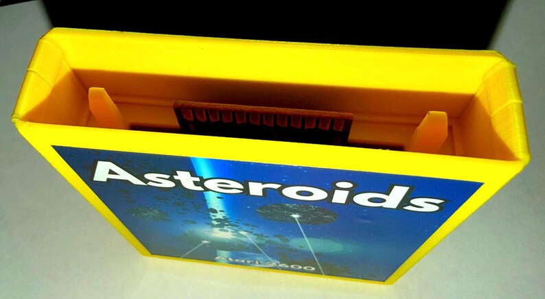 Peut inclure: Une cartouche de jeu vid&eacute;o Atari 2600 jaune vif pour le jeu Asteroids. L'&eacute;tiquette pr&eacute;sente le texte blanc Asteroids sur un fond spatial bleu fonc&eacute; avec des rochers flottants et un faisceau lumineux, avec Atari 2600 imprim&eacute; en dessous.