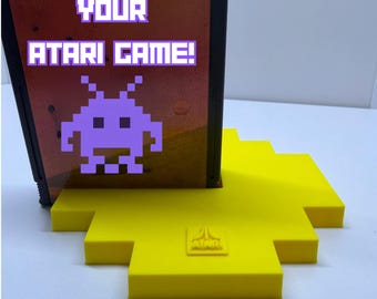 Peut inclure: Une cartouche de jeu Atari noire avec un extraterrestre pixelisé violet et le texte YOUR ATARI GAME! en violet, placée verticalement sur un support de présentation en plastique jaune vif en forme de croix avec le logo Atari en relief.