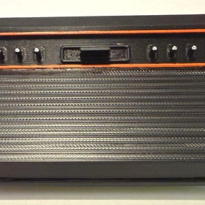 May include: A black rectangular electronic console featuring a ribbed front panel and a recessed top section outlined in orange. The top section contains six small vertical switches and one central rectangular slider switch, all in black.