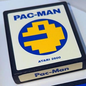 May include: A black rectangular Atari 2600 video game cartridge for Pac-Man. The label features a cream background with the blue text PAC-MAN at the top, a large yellow pixelated Pac-Man character inside a blue circle, and ATARI 2600 below.