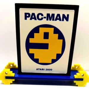 May include: A rectangular display sign featuring the text PAC-MAN and ATARI 2600 in blue on a white background. The center displays a yellow pixelated Pac-Man character inside a blue circle. The sign is supported by a blue base with yellow pixelated accents.