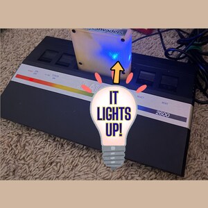 May include: A black Atari 2600 video game console with a silver control panel featuring a red power light and a game cartridge inserted in the top slot. The cartridge emits a bright blue light, and a lightbulb graphic displays the text IT LIGHTS UP!