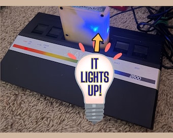 Peut inclure: Une console de jeu vidéo Atari 2600 noire avec un panneau de commande argenté, un voyant d'alimentation rouge et une cartouche insérée. La cartouche émet une lumière bleue vive, et un graphique d'ampoule affiche le texte IT LIGHTS UP!