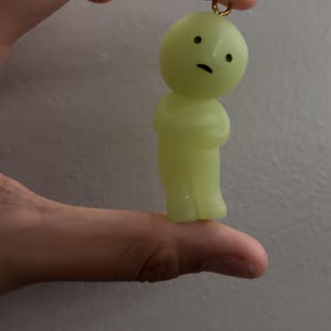 May include: A small, pale yellow, translucent figurine charm with a rounded head and a simple body. The figure features two black dots for eyes and a small black horizontal line for a mouth. A small gold-coloured metal loop is attached to the top.