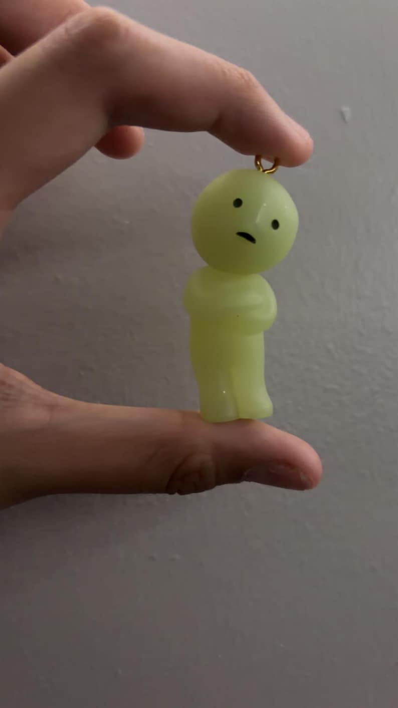 May include: A small, pale yellow, translucent figurine charm with a rounded head and a simple body. The figure features two black dots for eyes and a small black horizontal line for a mouth. A small gold-coloured metal loop is attached to the top.