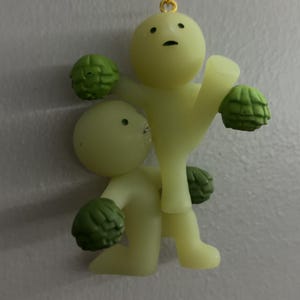 May include: A pale yellow keyring featuring two small, smooth figures with simple facial features. One figure stands while the other kneels, both holding textured green spherical objects resembling artichokes. A gold-toned metal chain is attached.